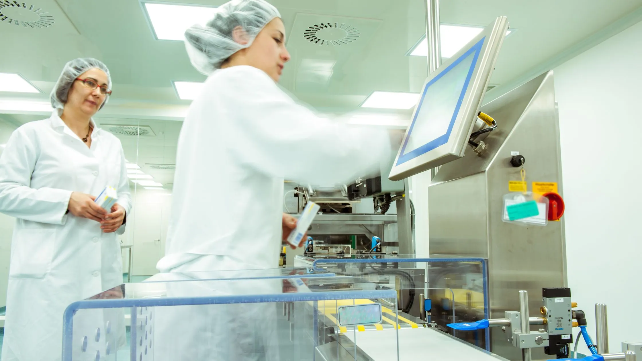 Two operators in cleanroom coats and hairnets running a pharmaceutical cartoning line