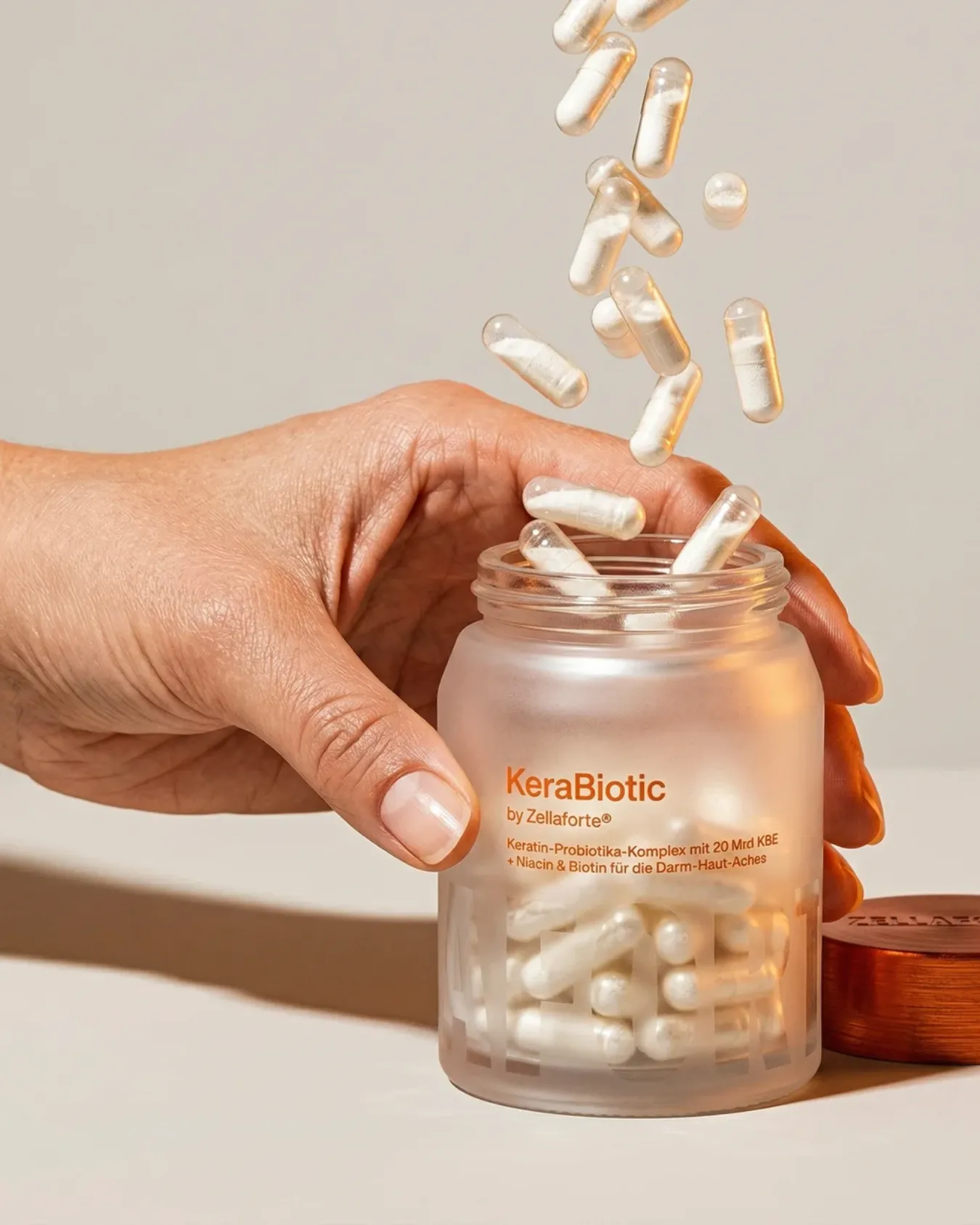 A hand holding the open Zellaforte® KeraBiotic jar as capsules fall in